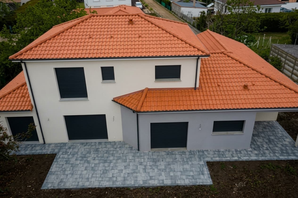 Maison moderne à deux étages, toit en tuiles orange, volets sombres et terrasse pavée grise.