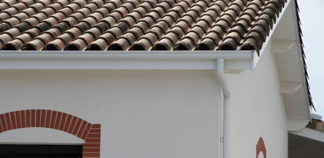 Gouttière blanche installée sous un toit en tuiles romanes sur une façade de maison blanche.