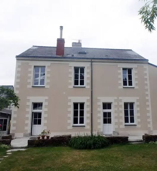 Maison beige à deux étages, fenêtres à encadrements blancs, toit en ardoise et cheminée.